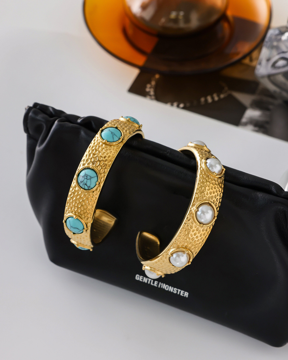 Gold hoop earrings with turquoise and pearl accents on a black pouch with 'Gentle Monster' branding.