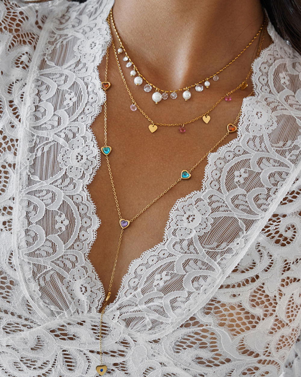 Layered gold necklaces with heart and teardrop pendants on a lace garment.