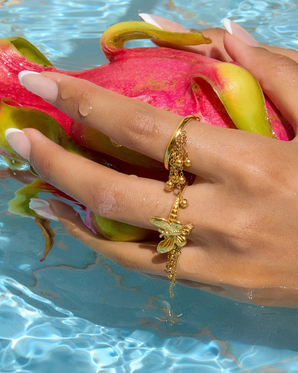 Gold ring with butterfly design worn on a hand holding a dragon fruit by a pool.