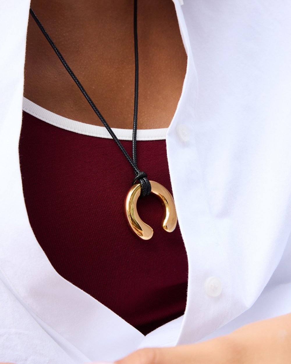 Gold pendant necklace on a black cord worn by a person with a white shirt and maroon top.