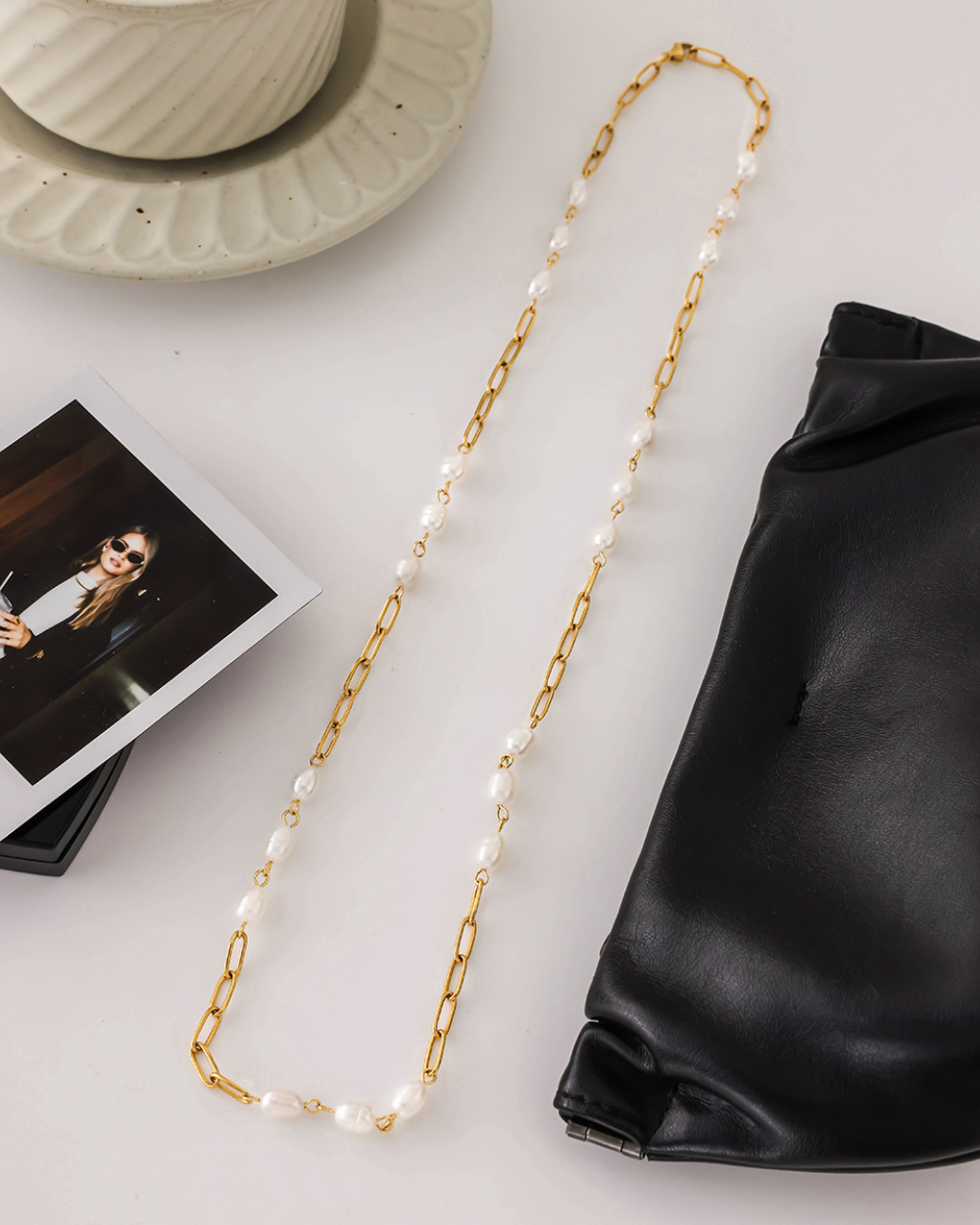 Gold chain with pearls on a white surface next to a black bag and photo album.