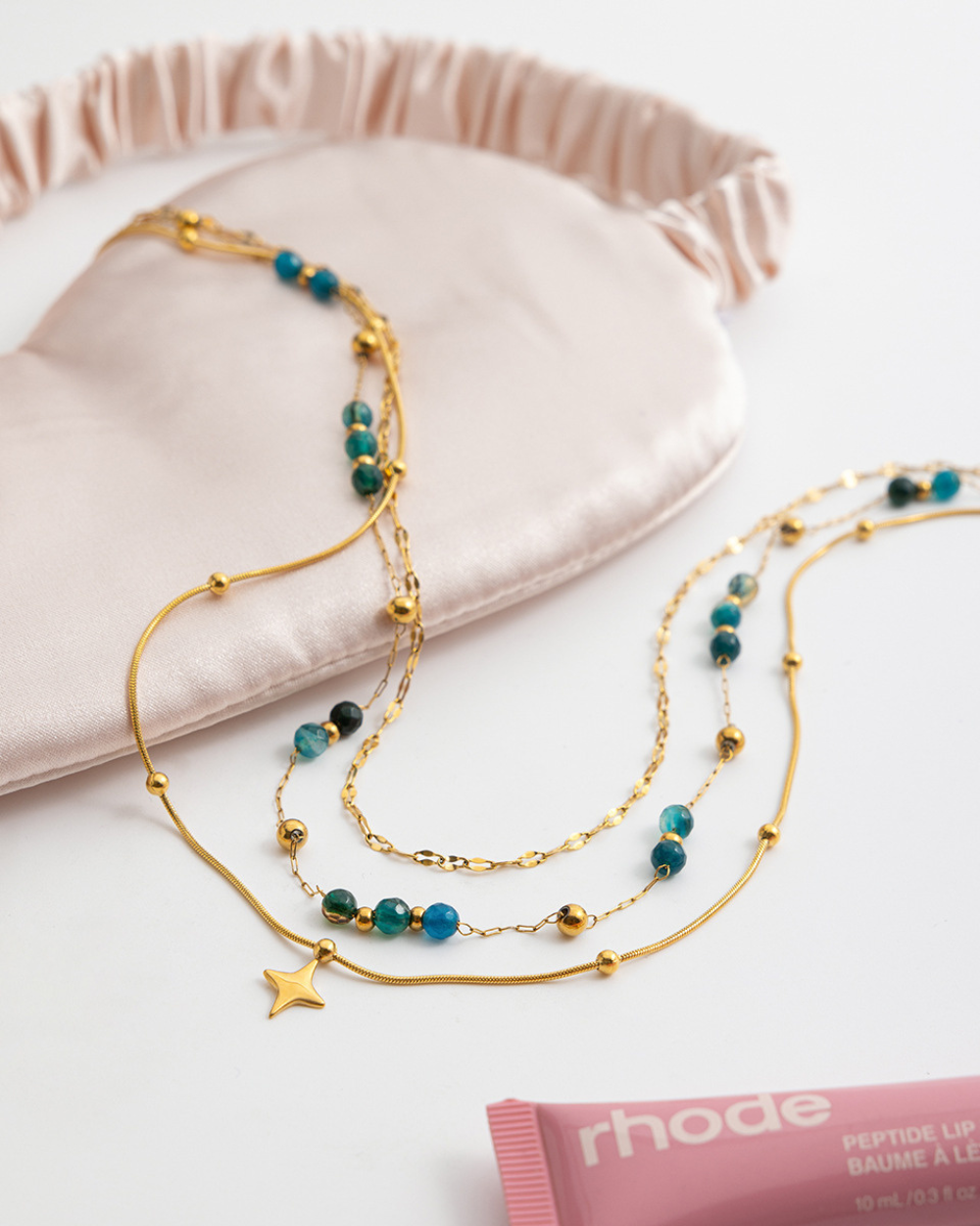 Gold and turquoise beaded necklaces on a pink surface with 'rhode' product in the background