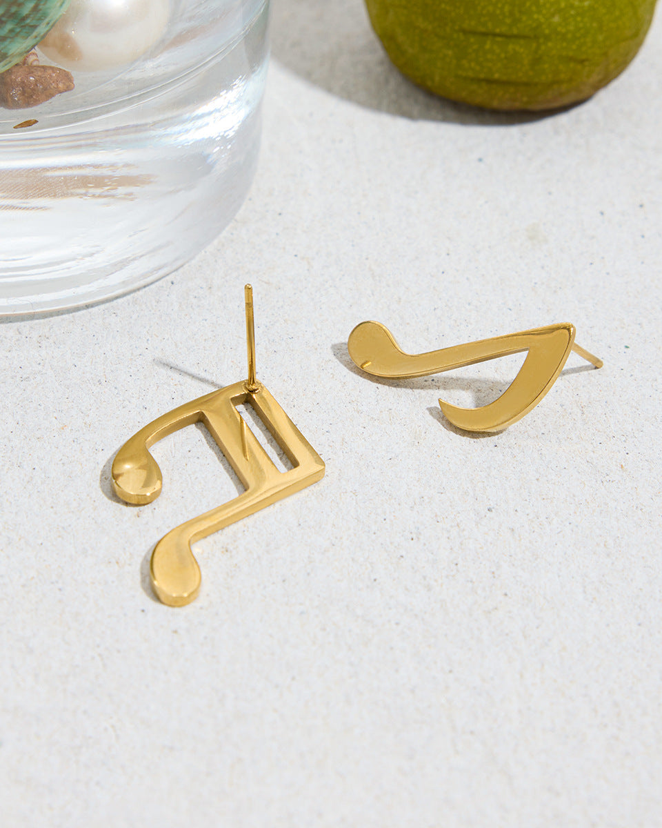 Gold music note earrings on a white surface with a glass of water and green object in the background.