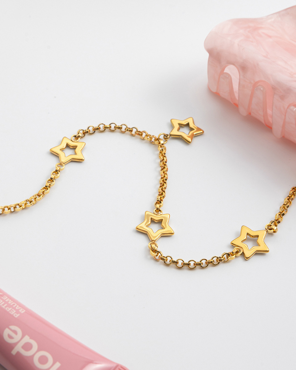 Gold star bracelet on a white surface with a pink tube of lip balm.