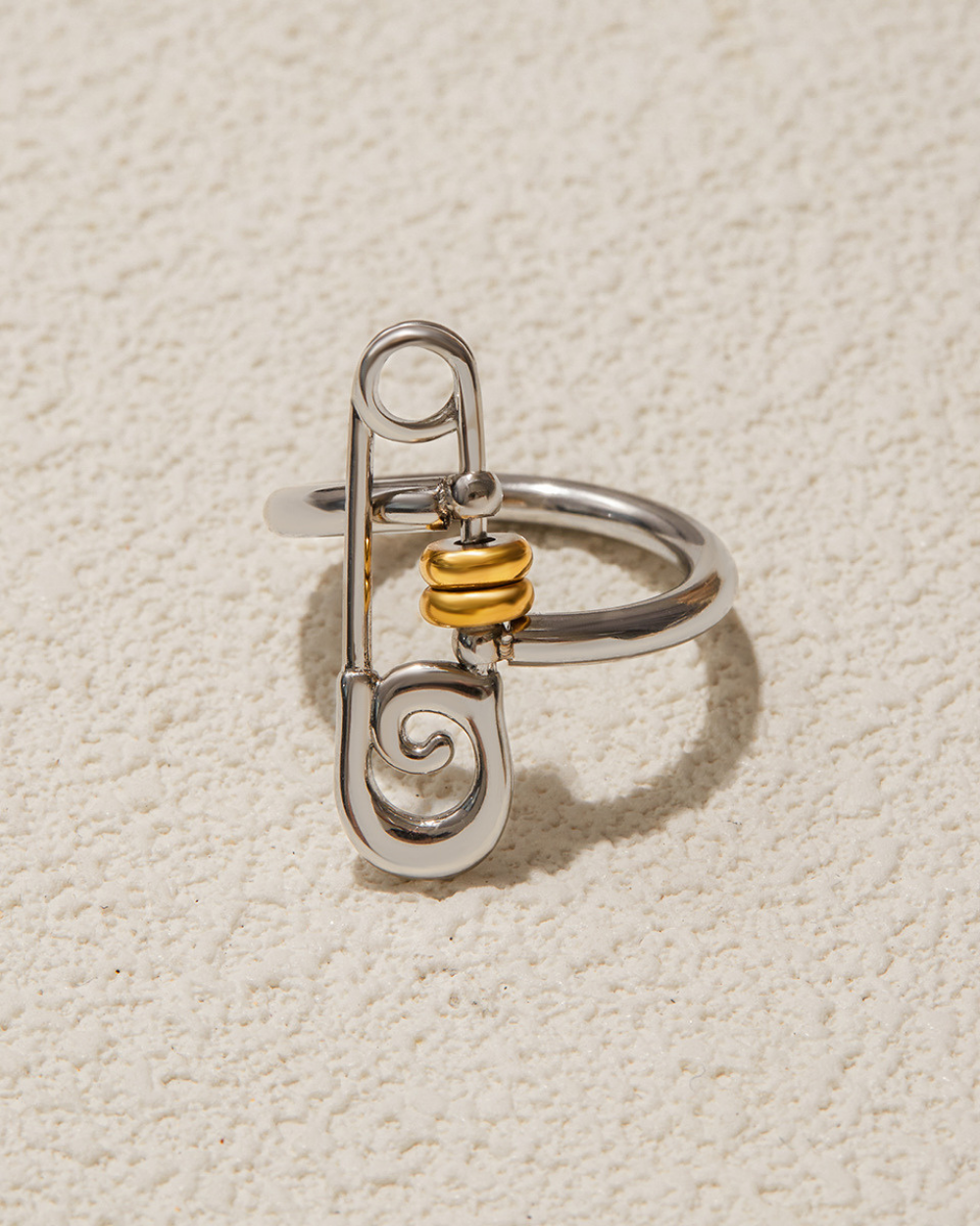 Silver safety pin-shaped ring with gold center on a beige textured background