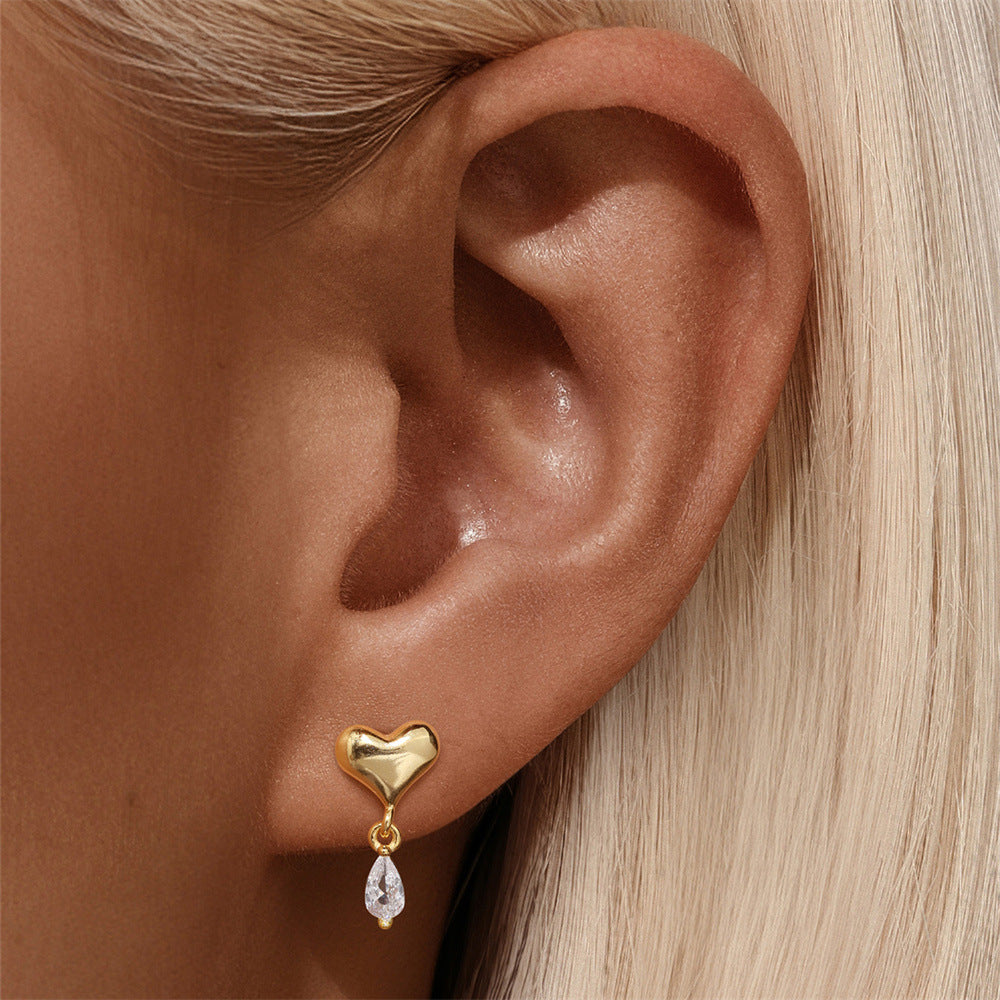 Pair of gold heart earrings with dangling pear-shaped crystal on white background