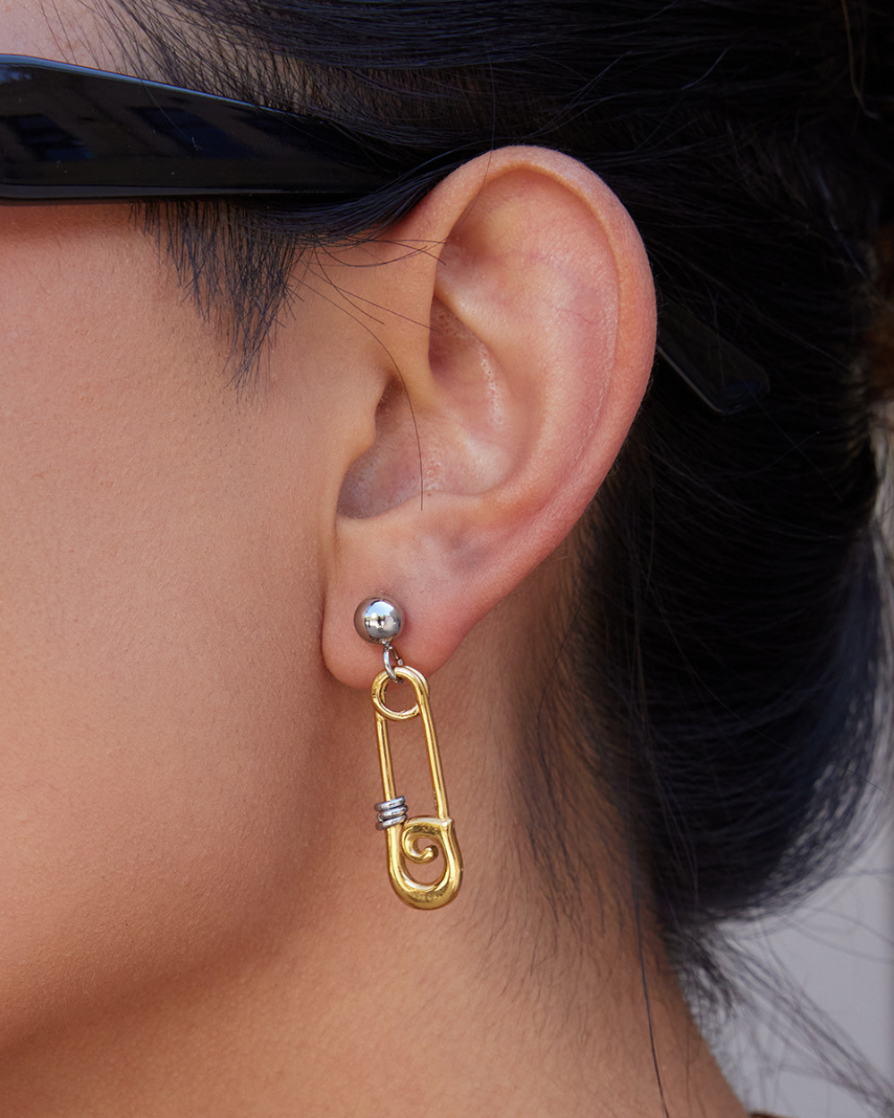 Gold safety pin earring worn by a person with sunglasses on.