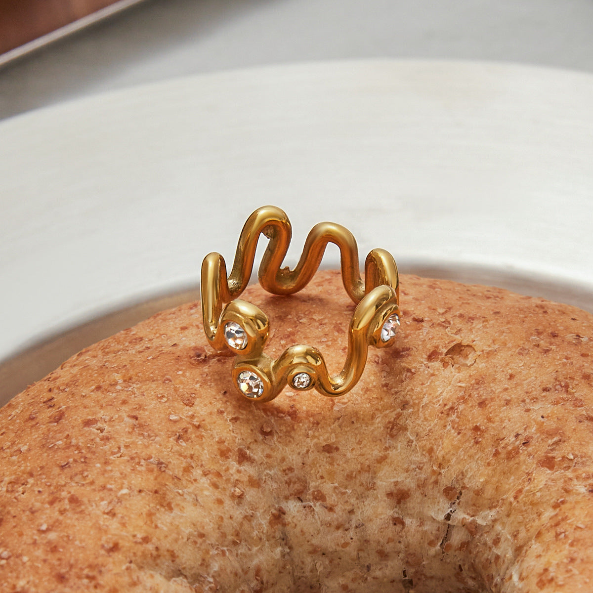 Gold crystal wave ring placed on a rustic bagel in a natural light setting