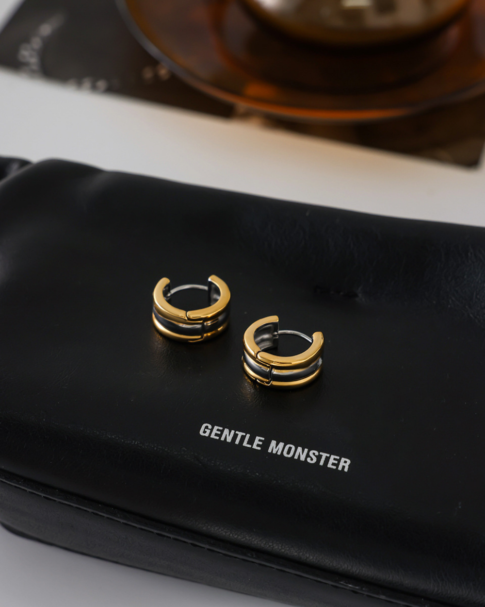 Gold hoop earrings on a black case with 'Gentle Monster' branding.