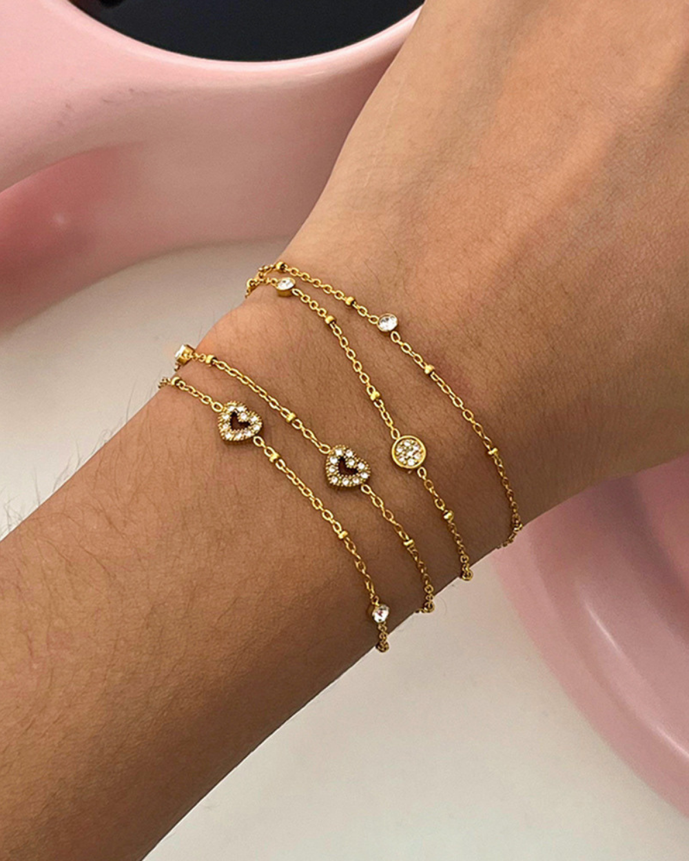 Gold bracelet with heart and flower designs on a wrist against a pink background