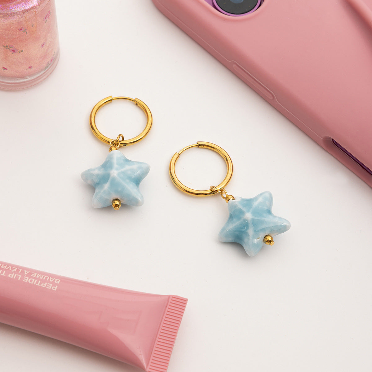 Star-shaped earrings with gold hooks on a white surface with pink phone and lip balm.