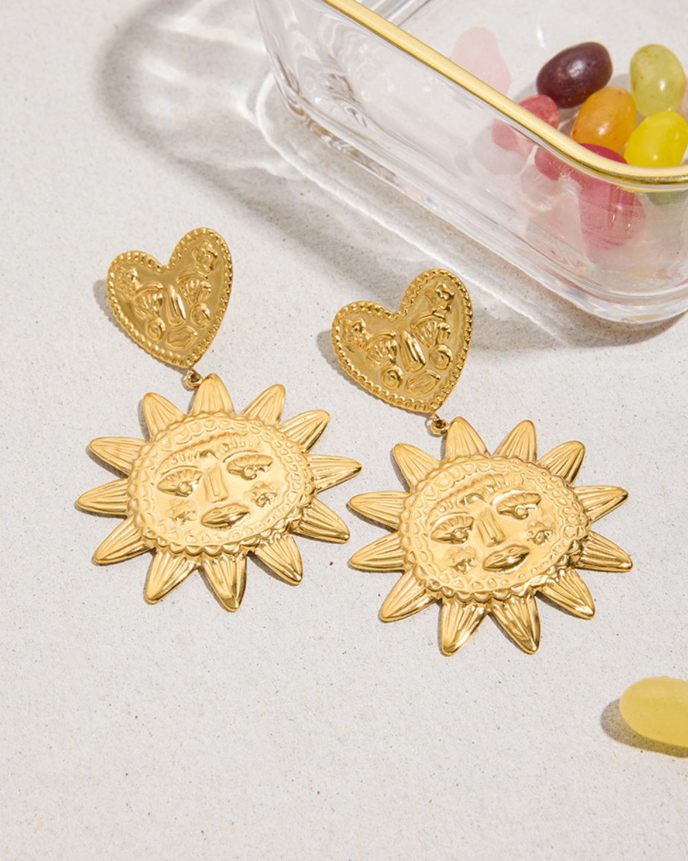 Gold sun-shaped earrings with heart pendants on a light surface.
