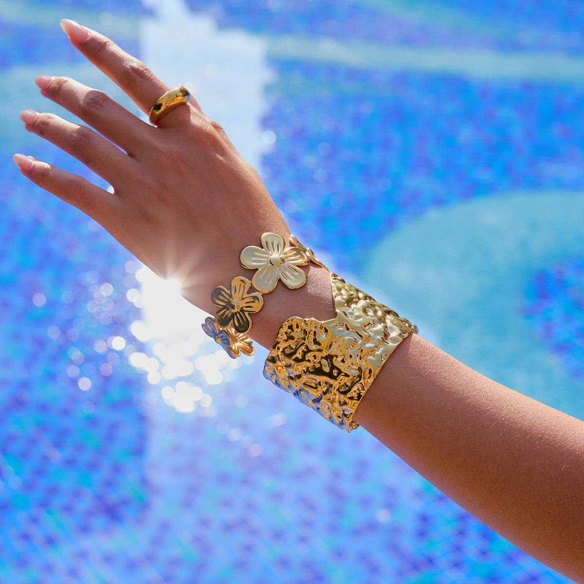 Hand reaching toward the sun wearing a floral gold cuff by the poolside