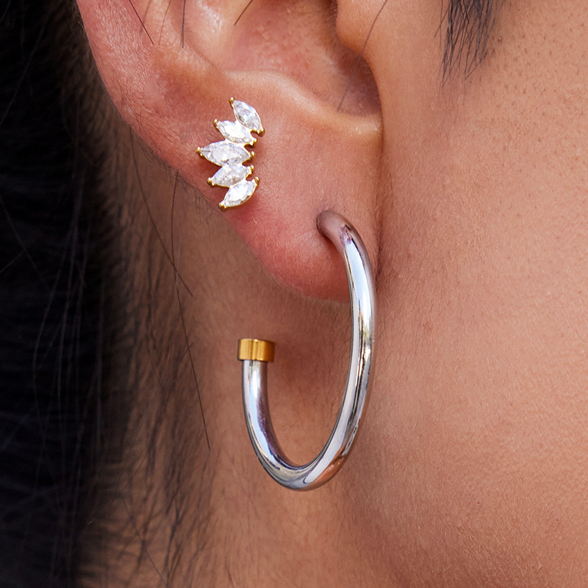 Close-up of an ear wearing a silver hoop earring with gold accent and a clear gemstone earring.
