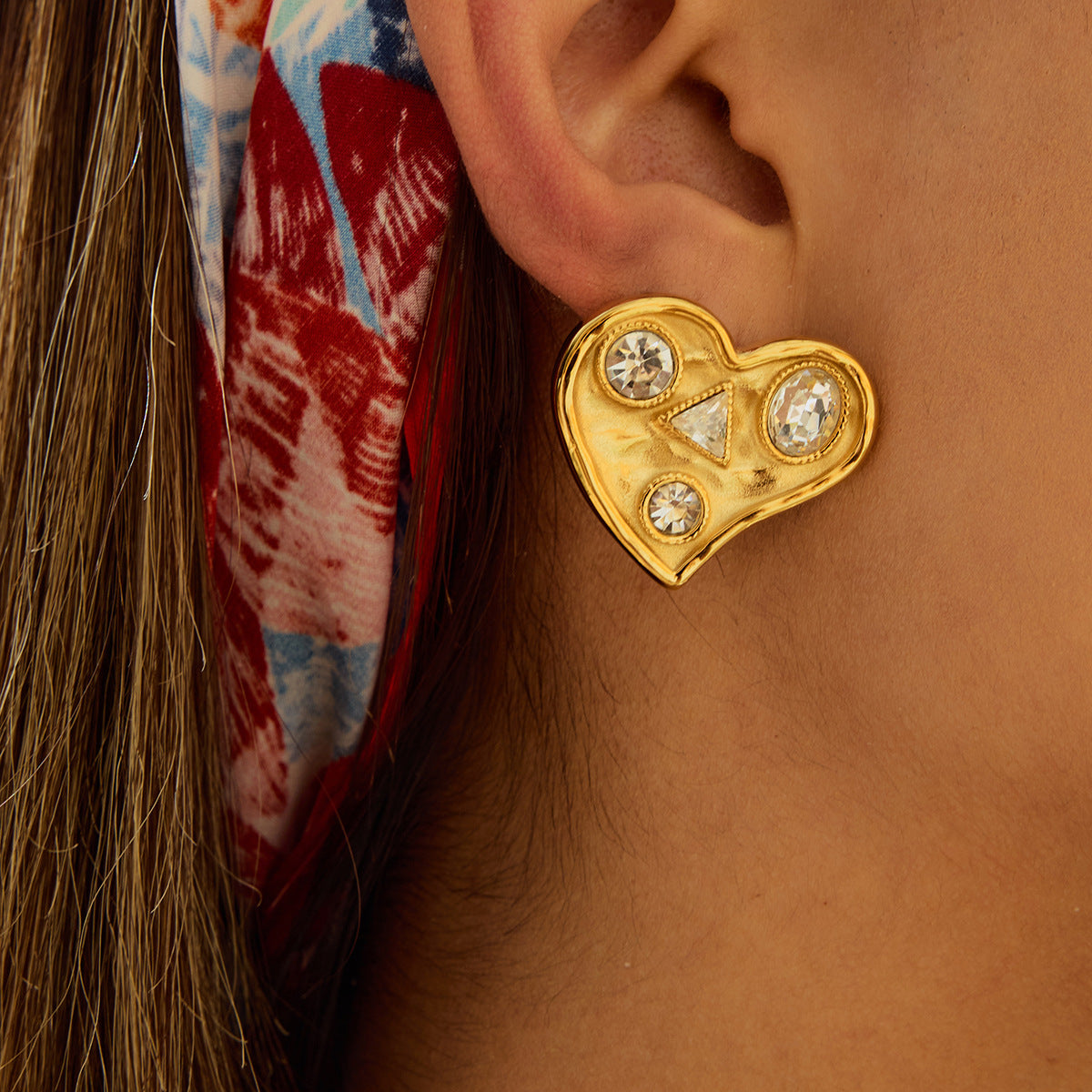 Model wearing large gold heart-shaped earrings with triangle and round stones, styled with colorful scarf.