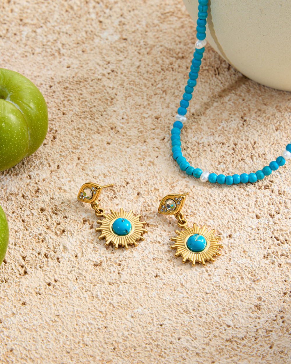 Gold earrings with turquoise stones and a blue beaded necklace on sand