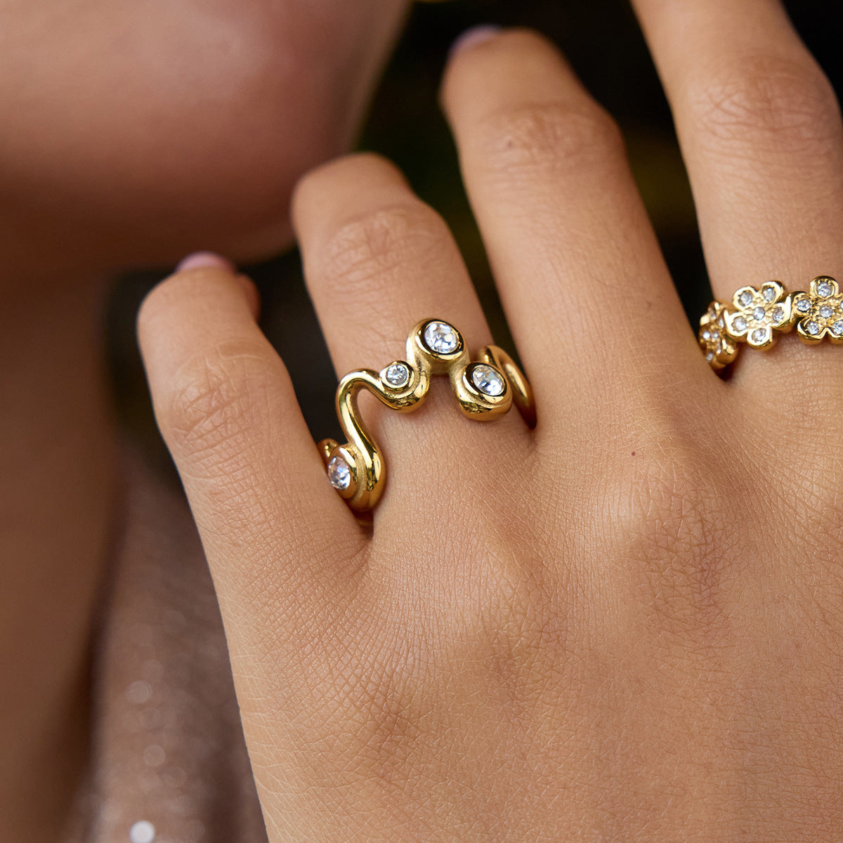 Model’s hand wearing gold wave ring with zircons and a floral ring, holding glass