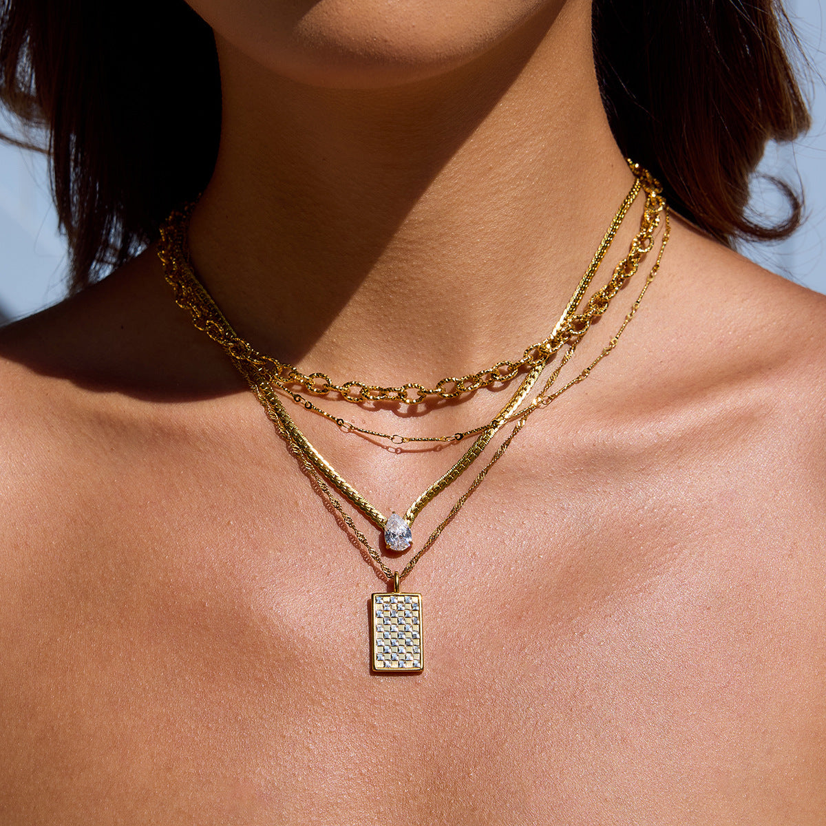 Model wearing layered gold necklaces featuring a square grid pendant in sunlight.