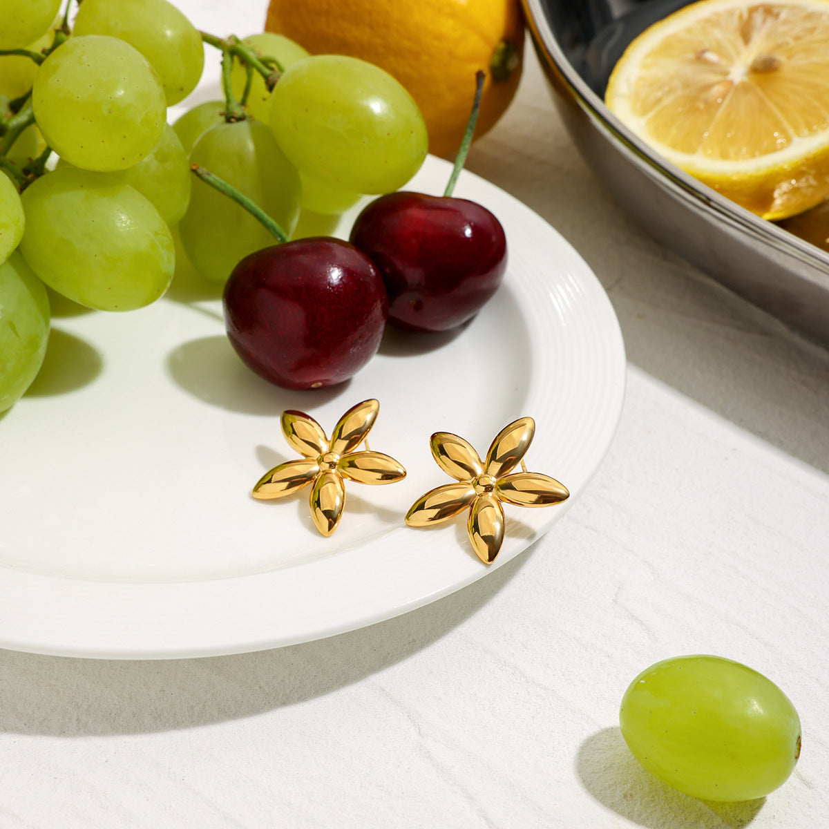 Pair of gold flower stud earrings on white surface with grapes