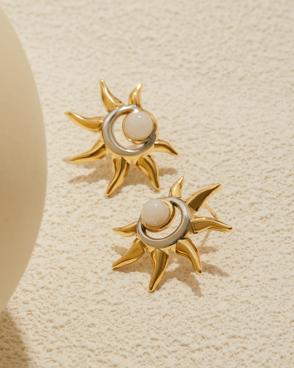 Gold sun-shaped earrings with a central pearl on a beige textured background