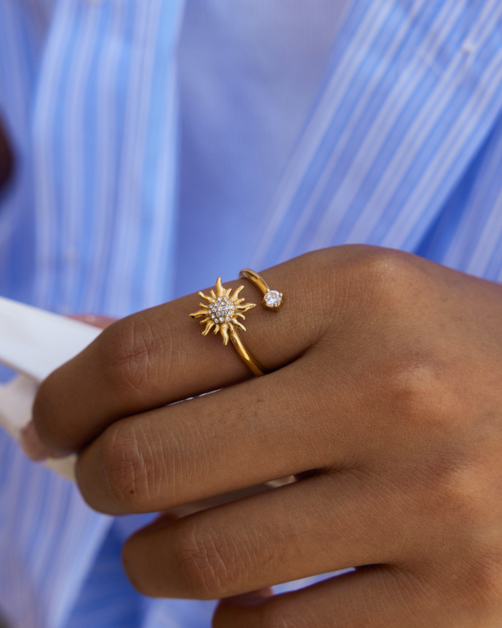 Hand wearing a gold ring with a sun design against a blue background