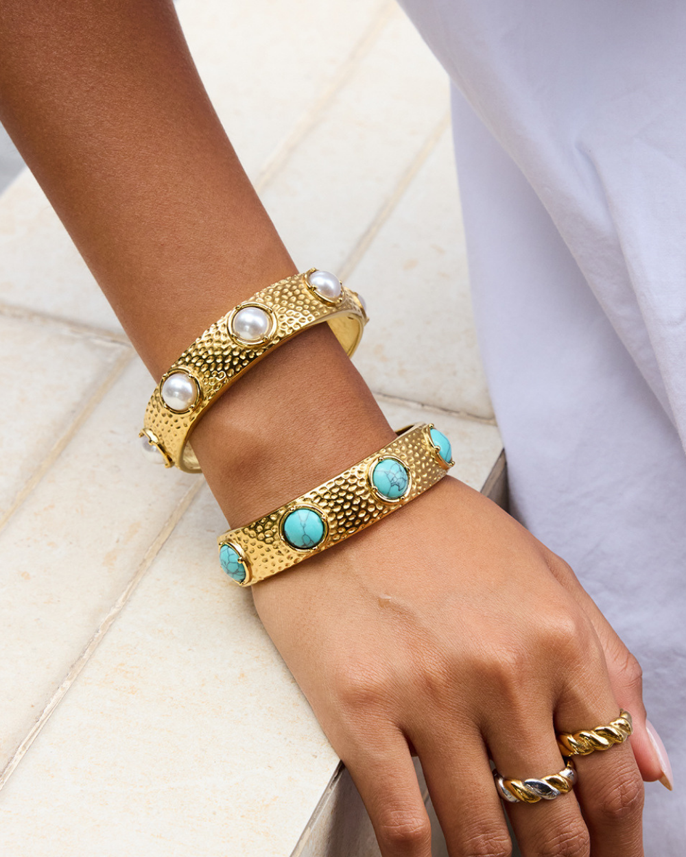 Gold bracelets with turquoise stones and pearls on a person's wrist against a white background