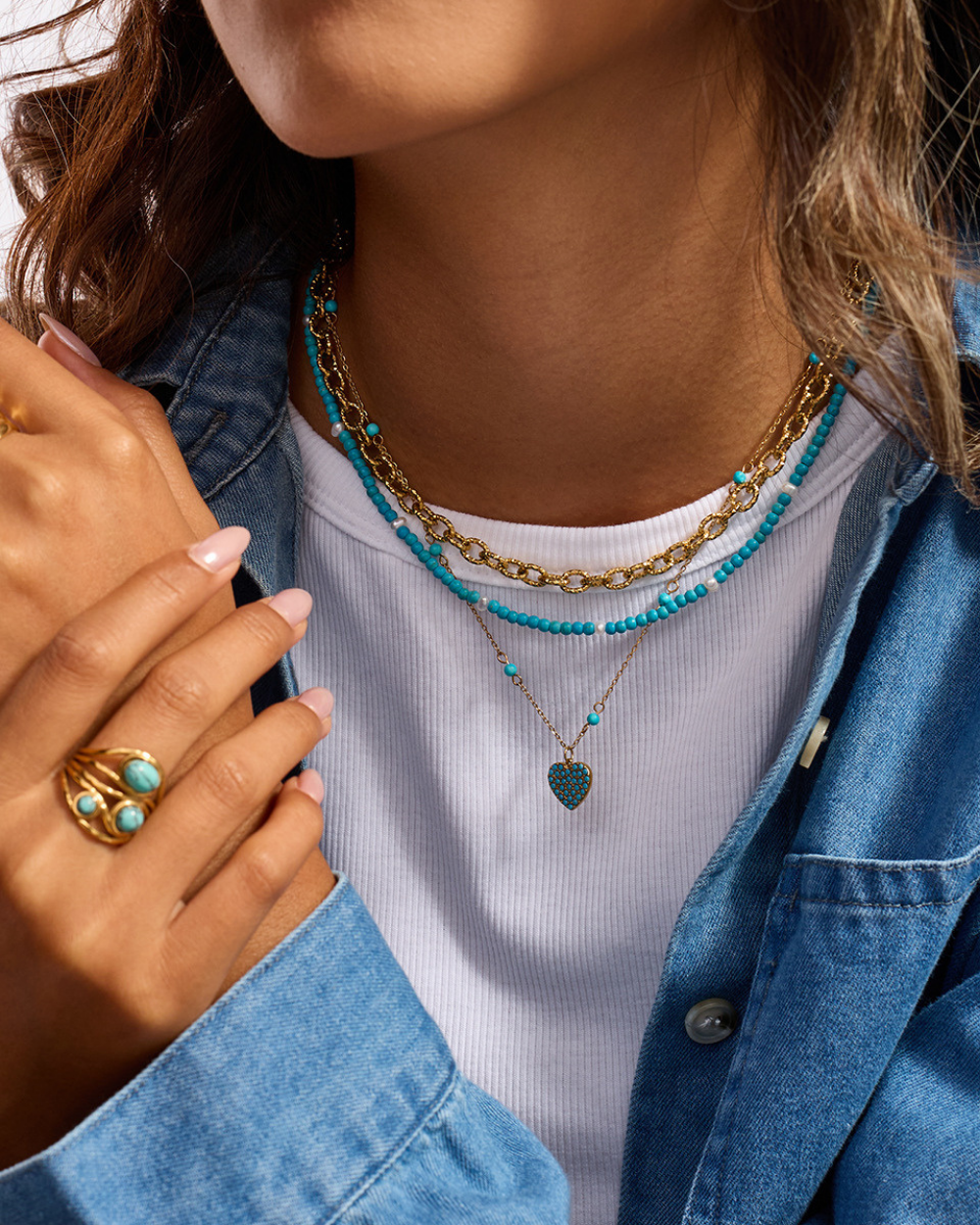 Person wearing multiple necklaces and a ring with turquoise stones, wearing a denim jacket.