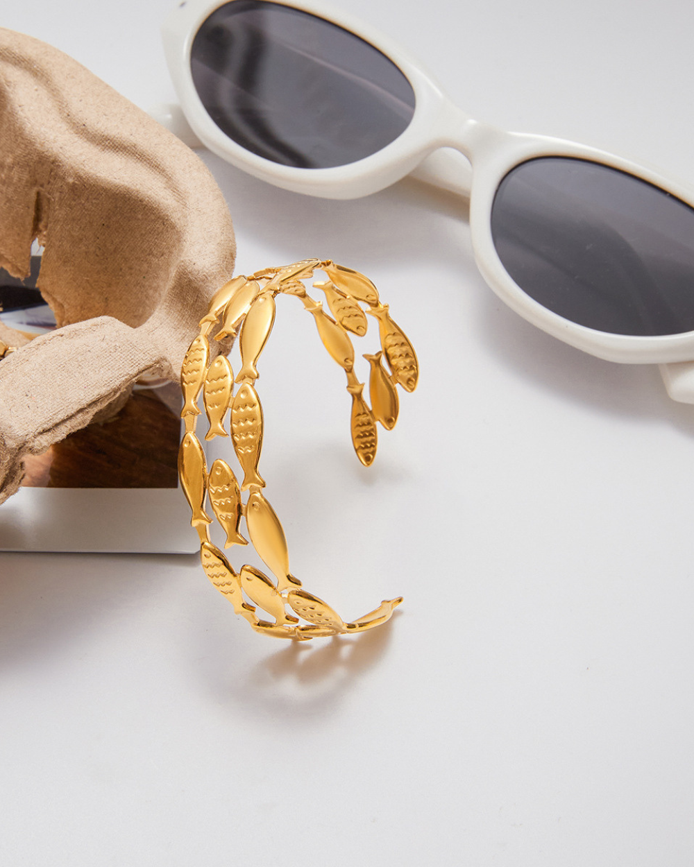 Gold bracelet with leaf design on a white surface next to sunglasses