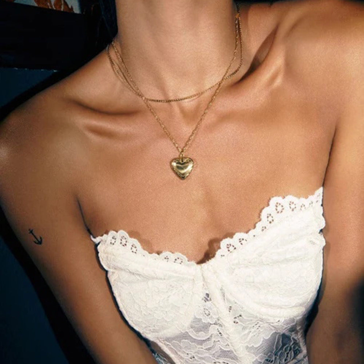 Close-up of a person wearing a gold necklace with a heart pendant and a white lace top.