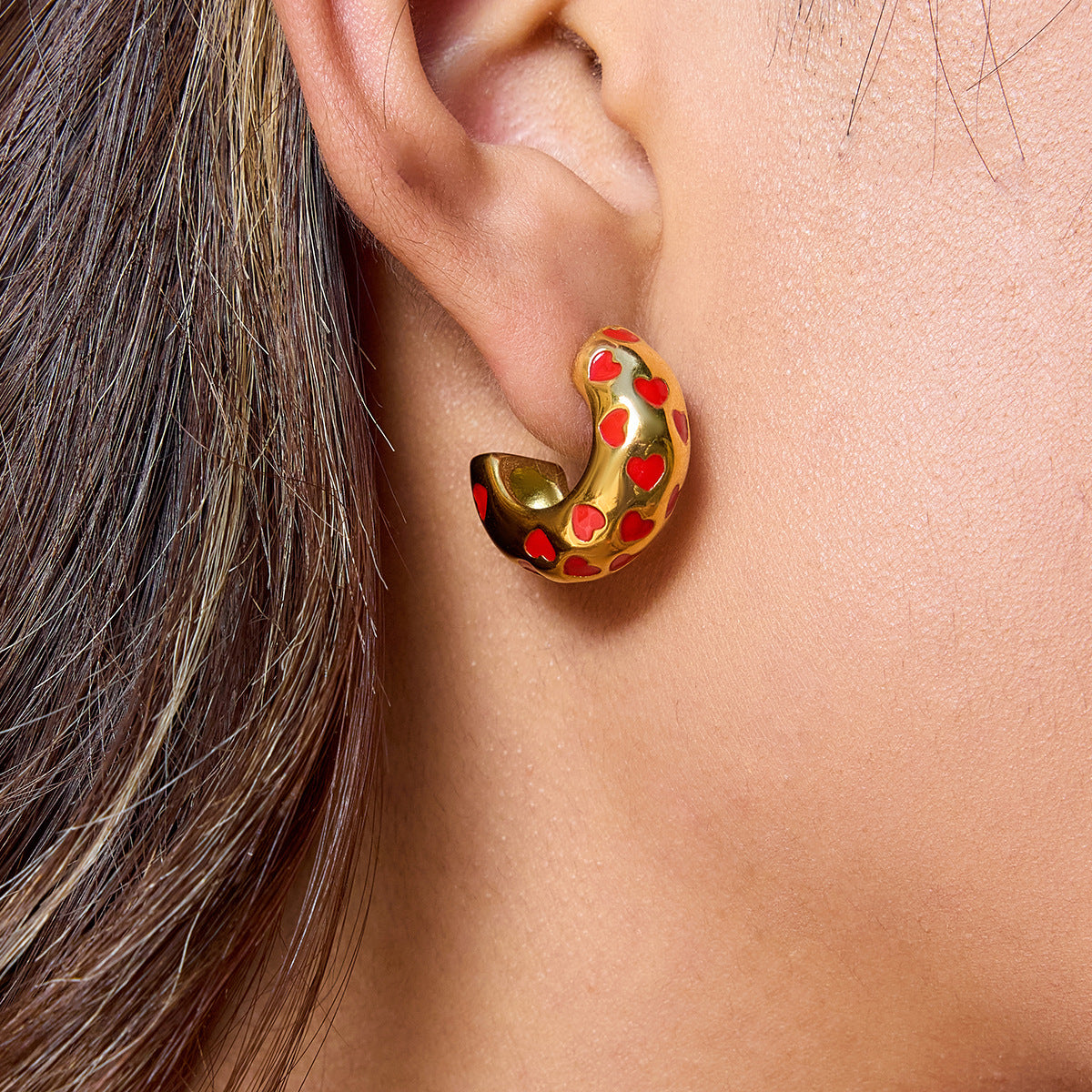 Model wearing bold gold heart earrings with glossy red enamel
