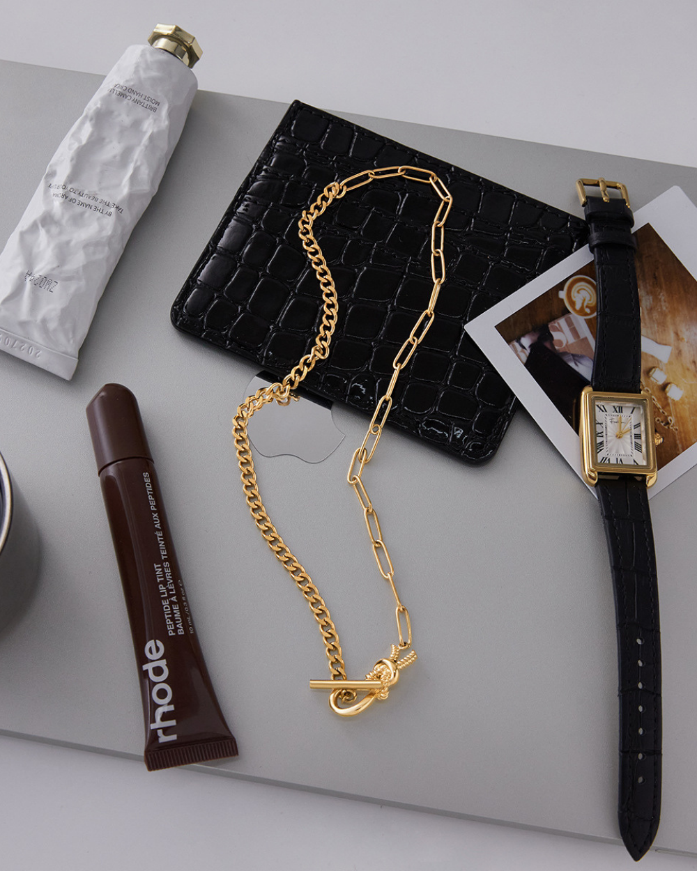 Gold chain necklace, black wallet, and watch on a light gray surface with a Rhode product.