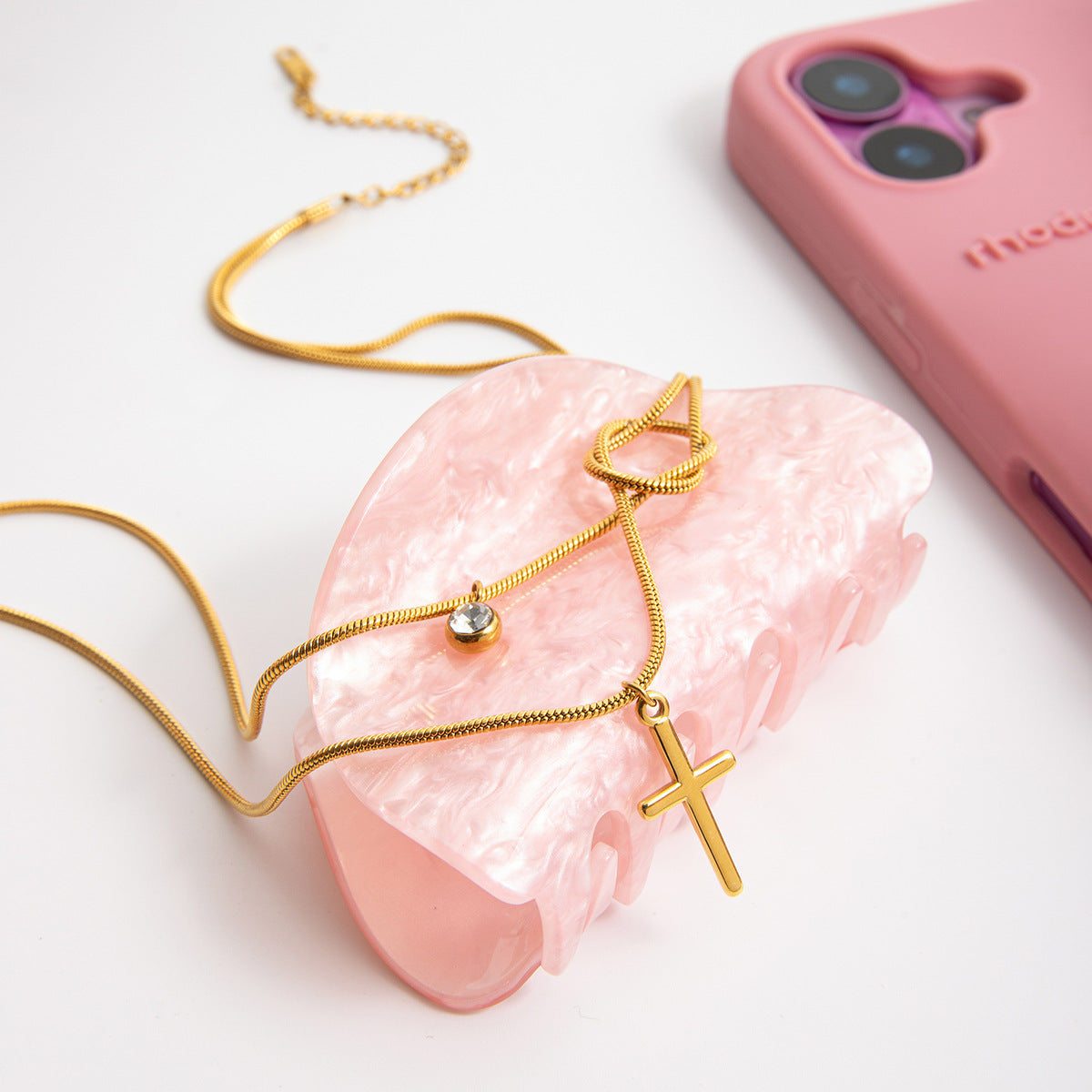 Gold necklace with a cross pendant on a pink stone, with a pink phone case in the background.