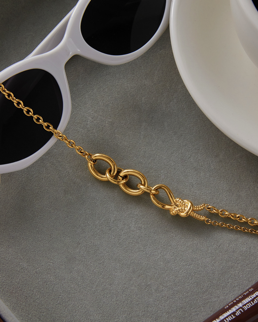 Gold chain necklace on a gray surface with sunglasses and a white dish in the background.