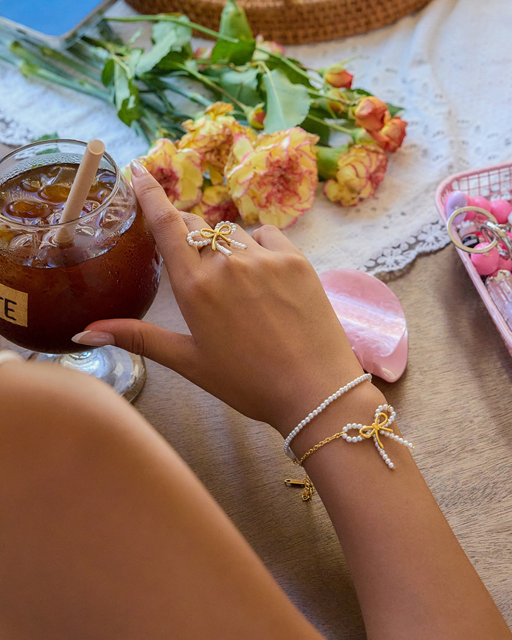 Hand with jewelry holding a drink, surrounded by flowers and a basket on a textured surface.