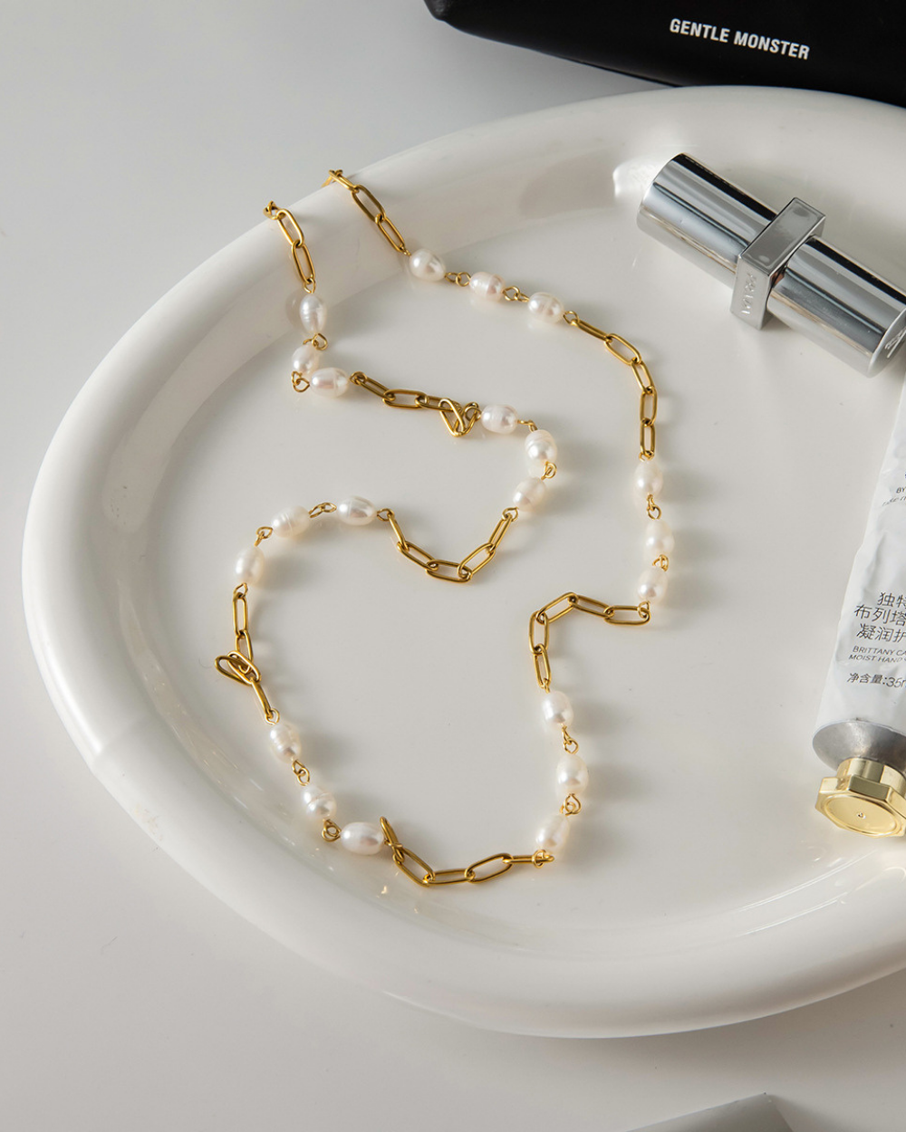 Pearl and gold chain necklace on a white plate with a bottle of skincare product.