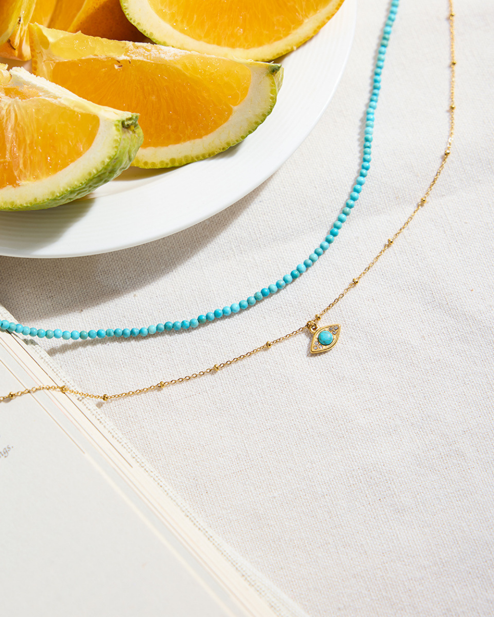 Turquoise beaded necklace with gold chain on a light surface with orange slices in the background