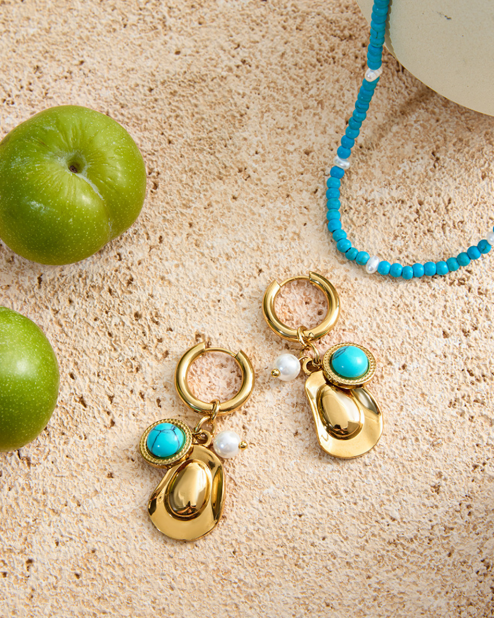 Gold earrings with turquoise stones and pearl accents on a textured surface with green apples and a blue beaded bracelet.