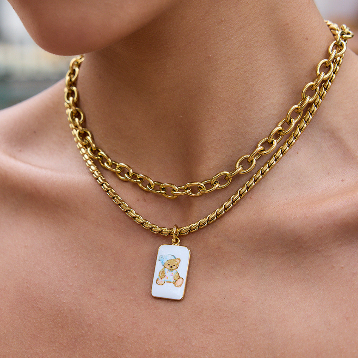 Model layering teddy bear necklace with bold chain, wearing floral dress outdoors