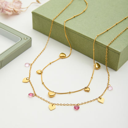 Gold necklace with heart and pink bead charms on a white surface with a green box in the background.