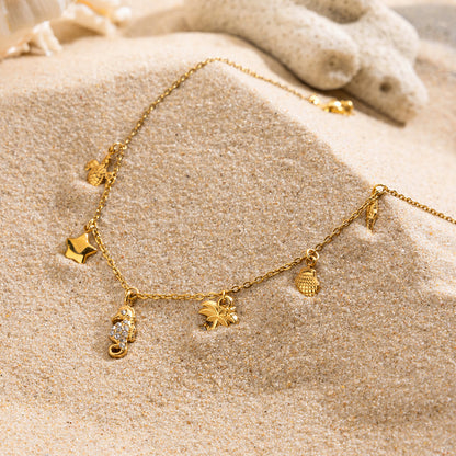 Flatlay of golden seahorse and palm tree charm bracelet on soft beach sand with seashells.