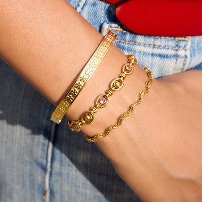 Close-up of a woman wearing three stacked gold bracelets, including a floral-engraved bangle, pink gemstone bracelet, and twisted rope chain, styled with denim and a red bag