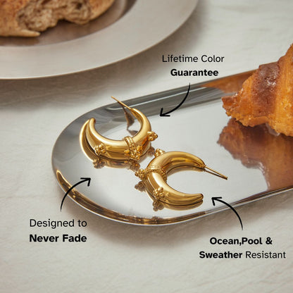 Crescent-shaped gold hoop earrings with ornate beaded detailing on reflective tray