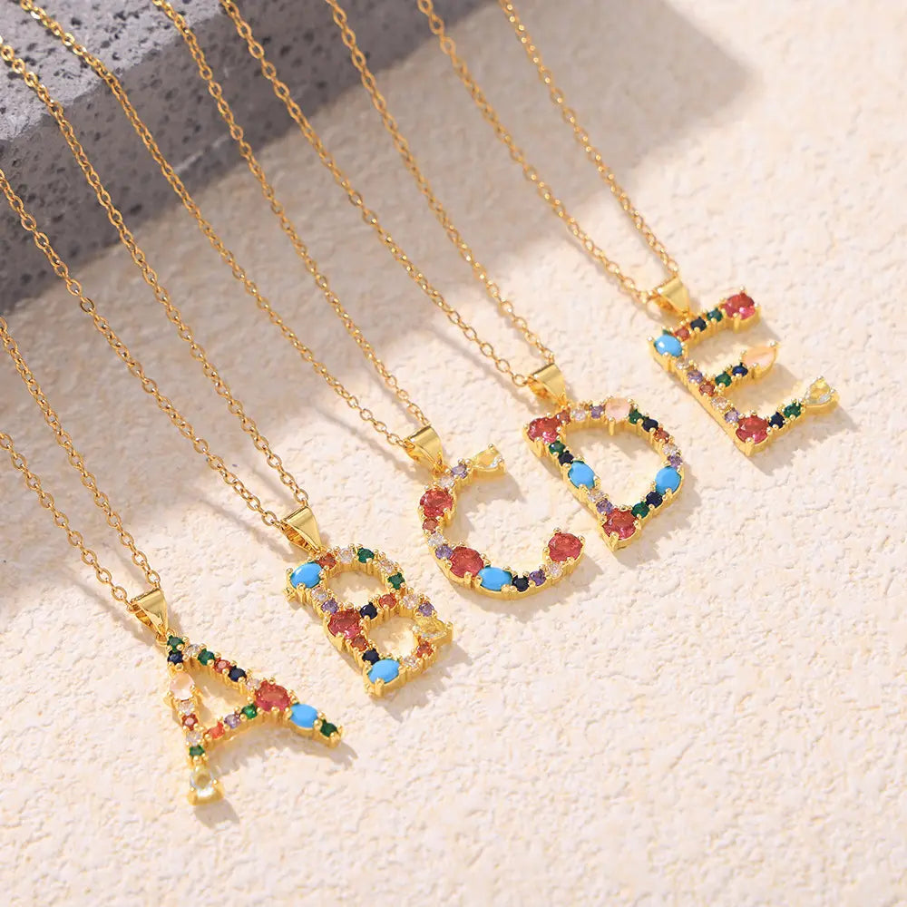 Colorful alphabet pendant necklaces featuring zircon stones in red, blue, green, and yellow, showcasing letters A, B, C, D, and E on a golden chain with a textured background.