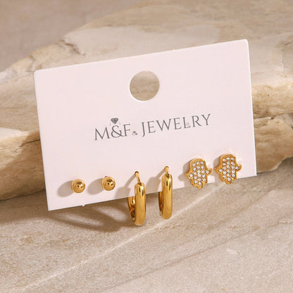 Gold-plated Hamsa hand earrings with clear crystals, laid flat on beige stone for lifestyle appeal.