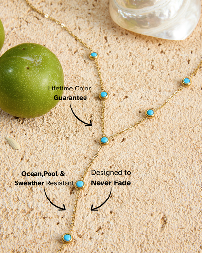 Gold necklace with turquoise stones on sand next to a green apple, with text about color guarantee and resistance.