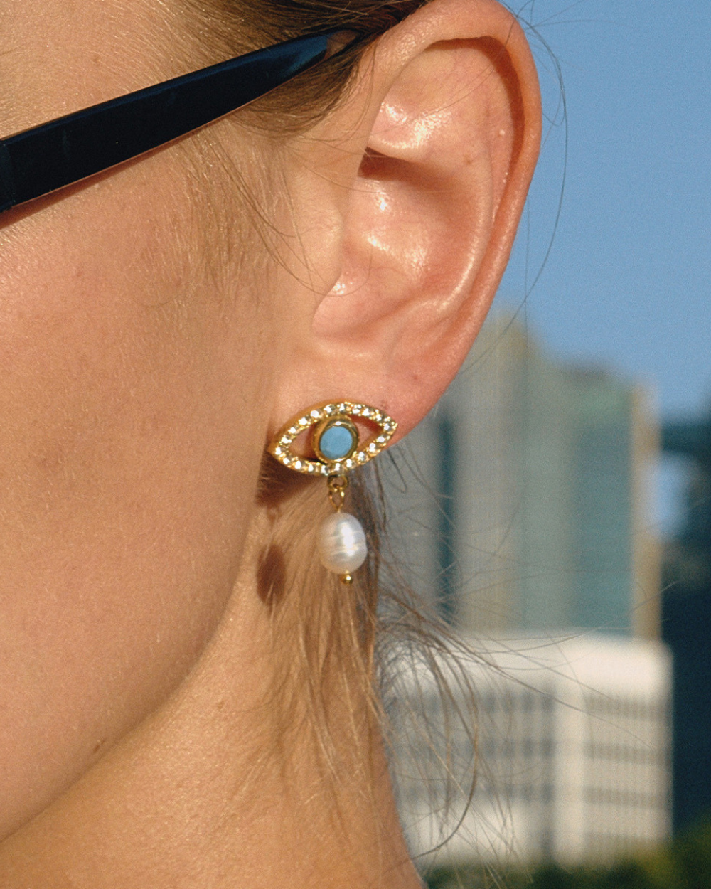 Model wearing evil eye pearl drop earrings with gold and turquoise detail, styled with sunglasses outdoors.
