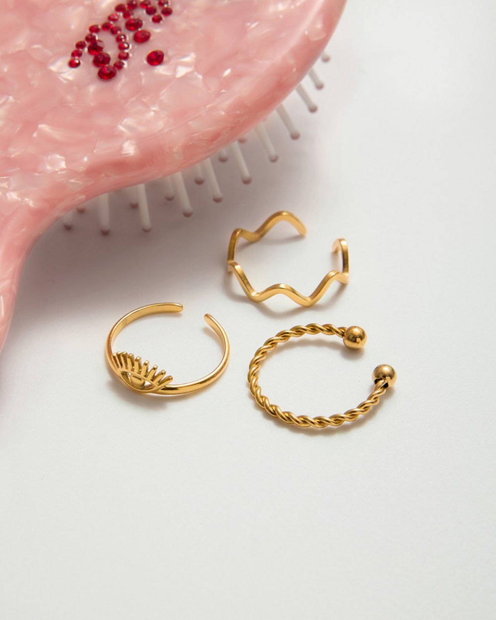 Flat lay on a white surface of the ring trio separated — open evil eye ring, zigzag band and twisted ring with ball ends — next to a pearly pink hairbrush.