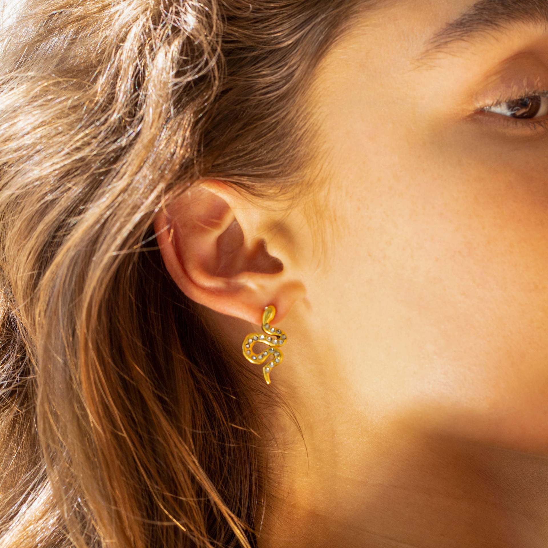Close-up of gold serpent earrings with crystals on model’s ear in sunlight