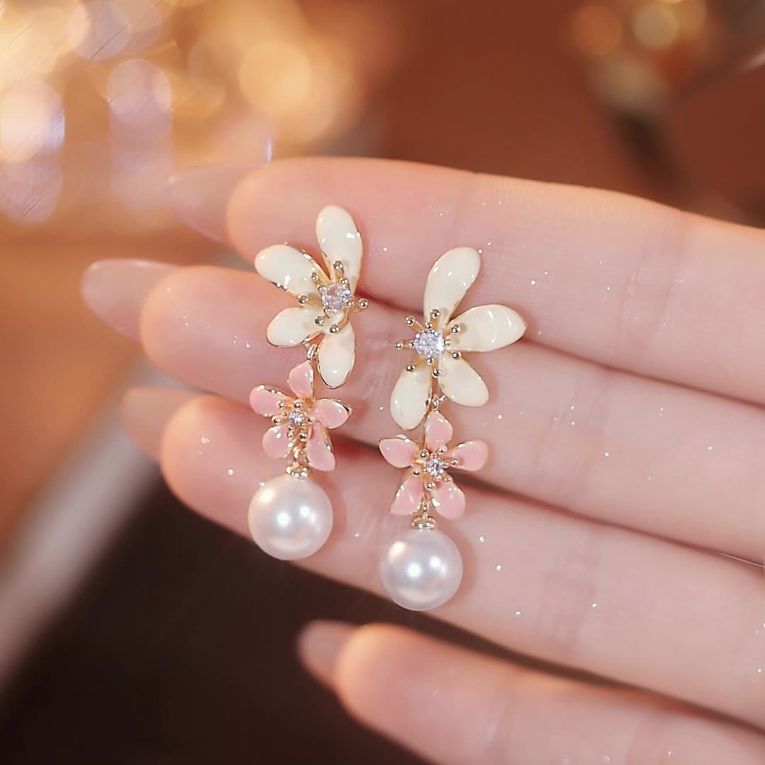 Delicate pearl flower drop earrings with pink and white petals, held by a woman with manicured nails in a soft beige setting.