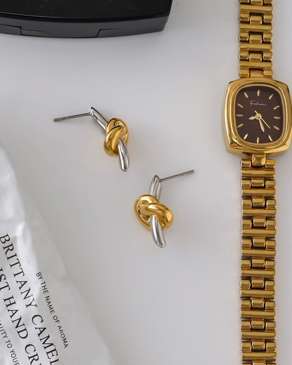Gold and silver knot earrings with a gold watch on a white surface.